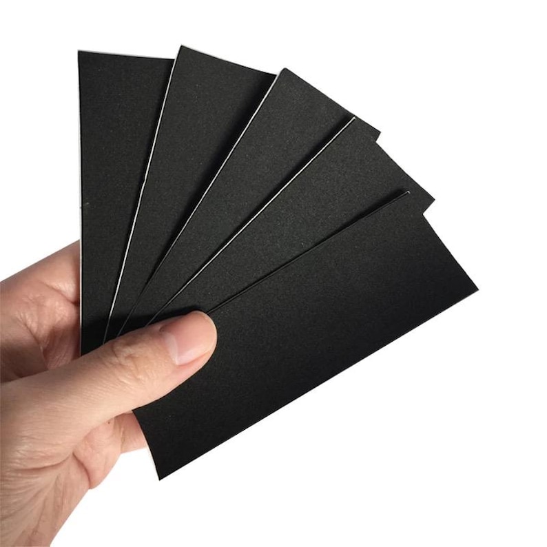 May include: A hand holding a fanned stack of rectangular black cards with white edges. The cards have a matte finish and are evenly spaced, creating a fan-like arrangement against a white background.