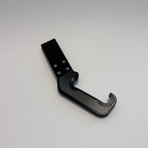 May include: A black plastic tool with a hook on one end and a flat, rectangular handle with four holes on the other end.