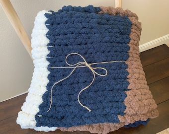 Könnte beinhalten: Eine dicke Strickdecke in Marineblau, Weiß und Braun. Die Decke ist mit Schnur gebunden und zur Hälfte gefaltet.