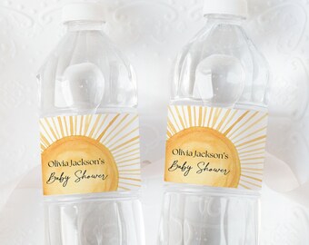 May include: Two clear plastic water bottles with yellow and orange sun-themed labels that say "Olivia Jackson's Baby Shower". The bottles are on a white plate with a decorative edge.