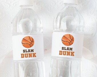 May include: Two clear plastic water bottles with white labels that say "Slam Dunk" and feature a basketball graphic. The bottles are on a white plate with a scalloped edge.