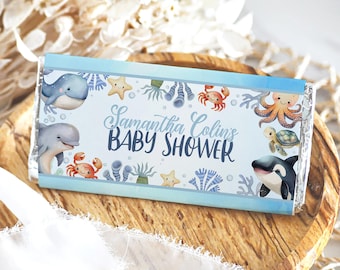 May include: A blue and white chocolate bar wrapper with a sea-themed design. It features various sea creatures like a whale, dolphin, octopus, crab, turtle, and starfish. The text on the wrapper reads "Samantha Colin's Baby Shower".
