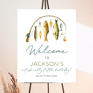 May include: A white welcome sign on an easel with a fishing theme. The sign features a watercolor illustration of a fishing rod with fish and lures. Text reads "Welcome to Jackson's a fish-ally fifth birthday!" and the date.