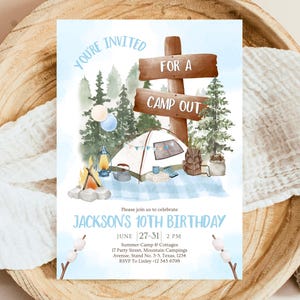 May include: A blue and white invitation for a camp out themed birthday party. The invitation features a cartoon illustration of a tent, a campfire, and a sign that says "For a Camp Out". The text on the invitation reads "You're Invited" and "Jackson's 10th Birthday".