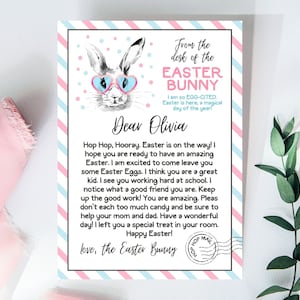 Puede incluir: Una tarjeta de rayas rosa y azul con un conejito de dibujos animados que lleva gafas de sol. La tarjeta tiene un mensaje del Conejo de Pascua para Olivia, deseándole una feliz Pascua y diciéndole que busque un regalo especial en su habitación.