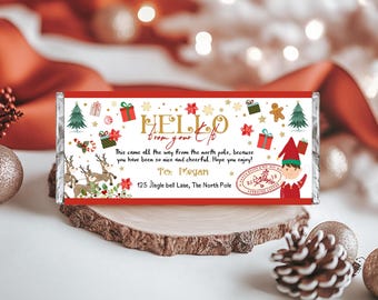 May include: A white chocolate bar wrapper with a red border. The wrapper features a festive Christmas design with an elf, reindeer, presents, candy canes, and stars. The text on the wrapper reads "HELLO from your Elf" and "This came all the way from the north pole, because you have been so nice and cheerful. Hope you enjoy! To: Megan 125 Jingle bell Lane, The North Pole".