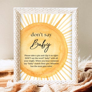 Könnte beinhalten: Ein gelbes und weißes Schild in Sonnenform mit dem Text "Sag nicht Baby" und Anweisungen für ein Spiel. Das Schild sagt, dass man eine Stecknadel nehmen und festklemmen soll, und während des Spiels das Wort "Baby" nicht sagen darf. Wer am Ende die meisten Stecknadeln hat, gewinnt.