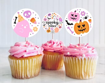 May include: Three pink frosted cupcakes with white sprinkles topped with Halloween themed cupcake toppers. The toppers feature a ghost, a pumpkin with hearts for eyes, and a pumpkin with bats and the text "spooky vibes".