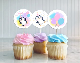 May include: Three cupcakes decorated with pink, purple, and blue frosting. Each cupcake has a round, white paper topper with a penguin design. One topper says "Let's Celebrate" with a penguin holding balloons. Another topper has a penguin wearing a party hat. The last topper has three balloons.