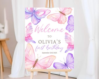 May include: A welcome sign for Olivia's first birthday, featuring watercolor butterflies in shades of pink and purple. The sign includes the text "Welcome to Olivia's first birthday" and the date.