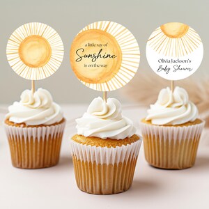 Könnte beinhalten: Drei Cupcakes mit weißem Frosting, verziert mit runden, gelben und weißen Cupcake-Toppern in Sonnenform. Die Topper zeigen den Text "a little ray of Sunshine is on the way" und "Olivia Jackson's Baby Shower".
