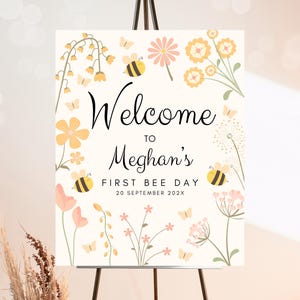 Op de afbeelding: Welkom op Meghans eerste bijendag, een wit bord met gele en roze bloemen, bijen en de tekst "Welkom op Meghans eerste bijendag 20 september 202X"