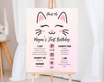 May include: Pink and white first birthday sign with a cute cat illustration. The sign includes the child's name, Megan, and details about their first year, including their weight, height, and favorite foods.