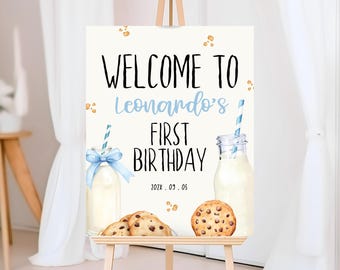 Op de afbeelding: Welkom bij Leonardo's First Birthday-bord met een blauw-wit gestreepte rietje, een glazen fles melk en twee chocoladekoekjes. Het bord staat op een houten ezel.