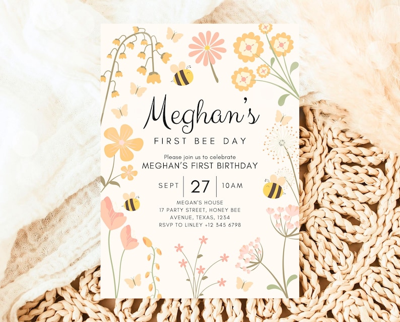 May include: A white invitation card with a floral design and a bee theme. The text on the card reads "Meghan's First Bee Day" and includes details for a first birthday party.