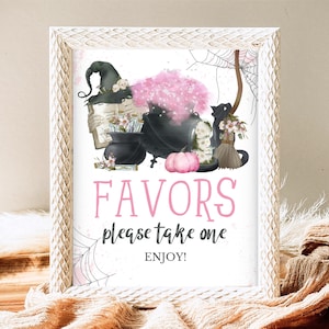 Op de afbeelding: Een wit frame met een roze en zwart Halloween-bord met de tekst "Favors please take one enjoy!" Het bord toont een heks, een ketel, een zwarte kat en een pompoen.