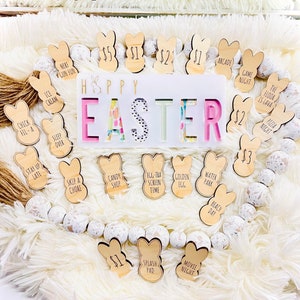 Puede incluir: Una decoraci&oacute;n de Pascua con etiquetas de madera en forma de conejito, cada una con una actividad o precio. La pieza central dice "HAPPY EASTER" con letras coloridas. La exhibici&oacute;n est&aacute; sobre un fondo blanco esponjoso con una guirnalda de cuentas.