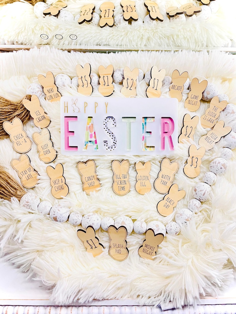 Puede incluir: Una decoraci&oacute;n de Pascua con etiquetas de madera en forma de conejito, cada una con una actividad o precio. La pieza central dice "HAPPY EASTER" con letras coloridas. La exhibici&oacute;n est&aacute; sobre un fondo blanco esponjoso con una guirnalda de cuentas.