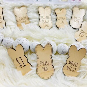Puede incluir: Etiquetas de madera con forma de conejito con frases como "Skip a Chore", "Candy Shop" y "Movie Night". Las etiquetas est&aacute;n dispuestas sobre una superficie blanca y esponjosa con esferas decorativas blancas y grises moteadas. Algunas etiquetas tienen precios.
