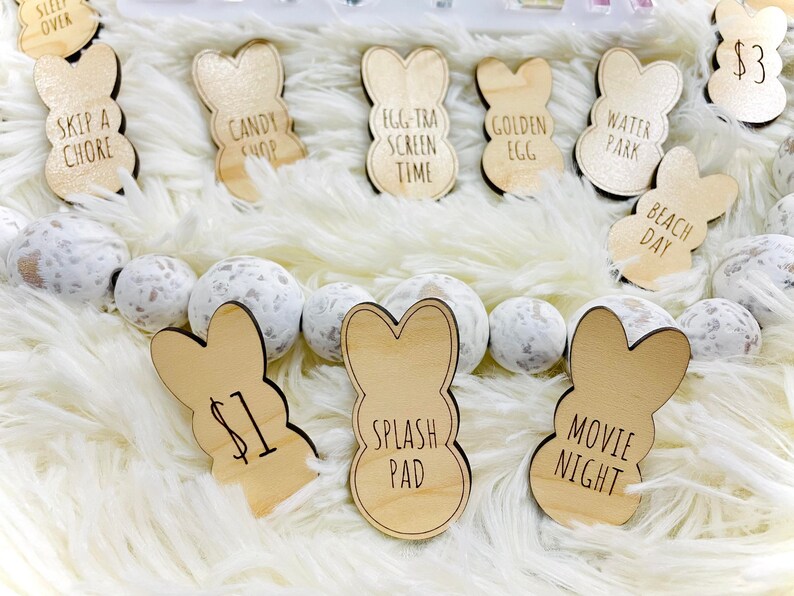 Puede incluir: Etiquetas de madera con forma de conejito con frases como "Skip a Chore", "Candy Shop" y "Movie Night". Las etiquetas est&aacute;n dispuestas sobre una superficie blanca y esponjosa con esferas decorativas blancas y grises moteadas. Algunas etiquetas tienen precios.