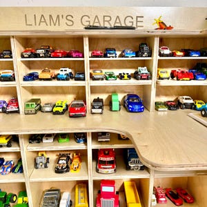 May include: A wooden display shelf labeled "LIAM'S GARAGE" filled with a collection of toy cars. The cars are various colors and styles, arranged in individual compartments. A small toy airplane sits on top.