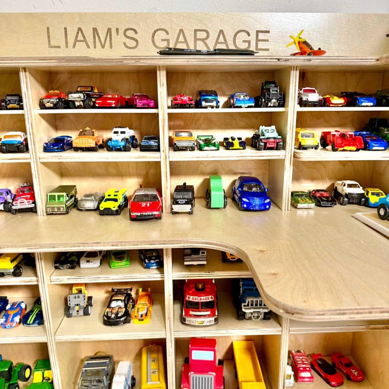 May include: A wooden display shelf labeled "LIAM'S GARAGE" filled with a collection of toy cars. The cars are various colors and styles, arranged in individual compartments. A small toy airplane sits on top.