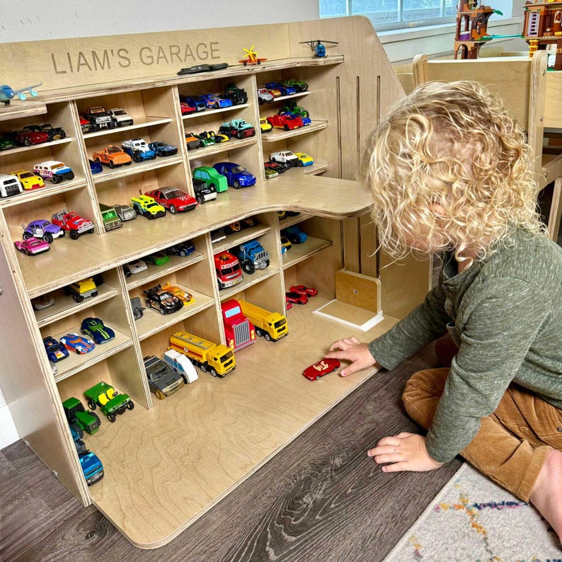 May include: A wooden toy garage with multiple shelves filled with colorful toy cars. The top of the garage is labeled "LIAM'S GARAGE". A child is playing with a red toy car in front of the garage.