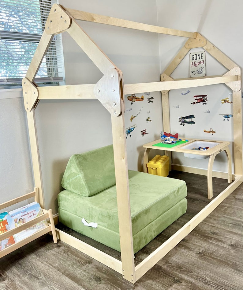 May include: A wooden playhouse frame with a green cushioned seat inside. The playhouse has a sign that says "Gone Flying".