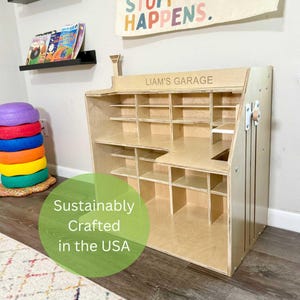 May include: A light-colored wooden storage unit with multiple shelves and compartments. The text "LIAM'S GARAGE" is visible on the top. A banner above reads "STUFF HAPPENS." A green circle states "Sustainably Crafted in the USA."