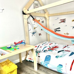May include: A wooden bed frame shaped like a house with a hammock hanging from the top. The bed has a white and blue airplane themed bedding and a yellow toy storage bin on the floor. The wall has airplane decals and a sign that says "Gone Flying".