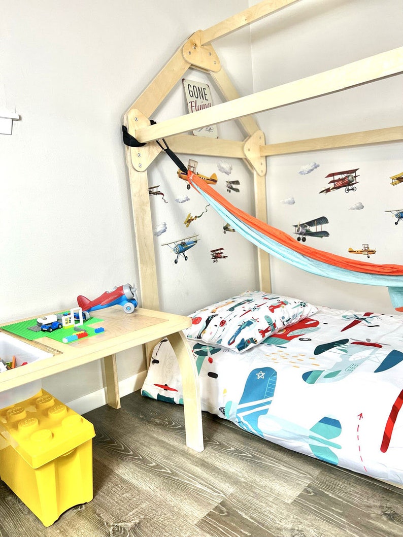 May include: A wooden bed frame shaped like a house with a hammock hanging from the top. The bed has a white and blue airplane themed bedding and a yellow toy storage bin on the floor. The wall has airplane decals and a sign that says "Gone Flying".