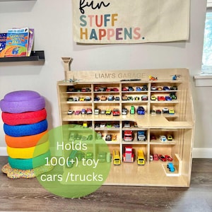 May include: A wooden toy car garage with multiple shelves filled with colorful toy cars and trucks. The text "LIAM'S GARAGE" is above the shelves. The text "Holds 100(+) toy cars / trucks" is in the foreground.