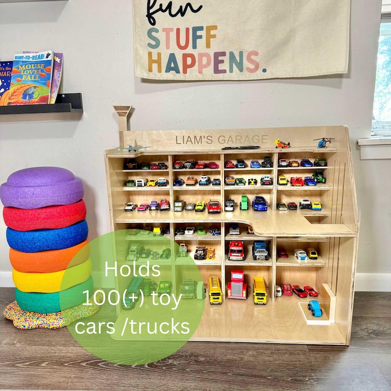 May include: A wooden toy car garage with multiple shelves filled with colorful toy cars and trucks. The text "LIAM'S GARAGE" is above the shelves. The text "Holds 100(+) toy cars / trucks" is in the foreground.
