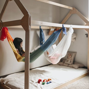 May include: A girl sits in a rainbow hammock reading a book inside a wooden playhouse. The playhouse has a white mattress and a set of colorful wooden rainbow arches on the floor.