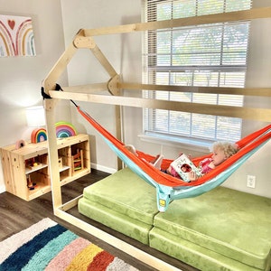 May include: A wooden playhouse frame with a hammock inside. A child is sitting in the hammock reading a book. The playhouse is in a room with a rainbow rug and a wooden shelf with toys.