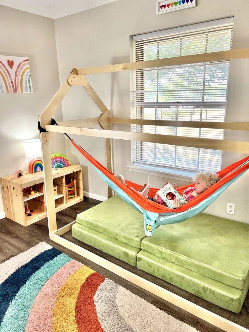May include: A wooden playhouse frame with a hammock inside. A child is sitting in the hammock reading a book. The playhouse is in a room with a rainbow rug and a wooden shelf with toys.