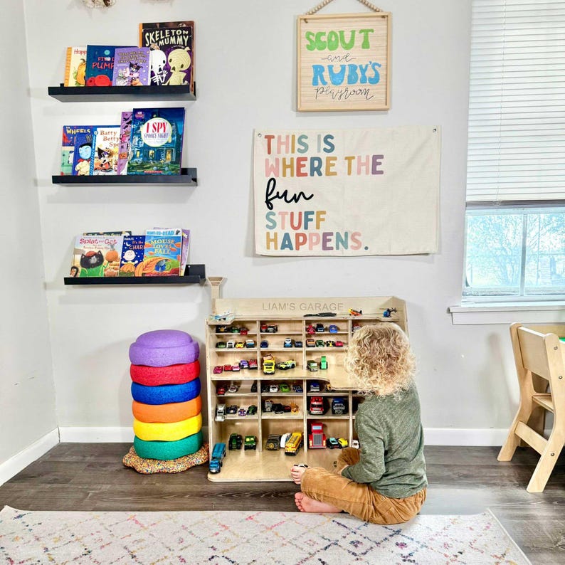 May include: A child's playroom with a wooden toy car display cabinet filled with various toy cars. The room features a stack of colorful cushions, bookshelves with children's books, and wall art with the text "THIS IS WHERE THE fun STUFF HAPPENS."