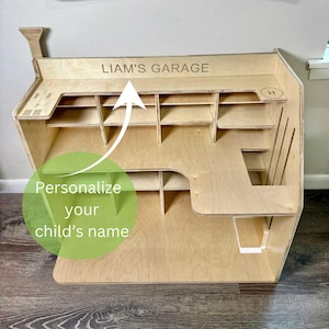 May include: A wooden toy garage with multiple shelves and compartments. The text "LIAM'S GARAGE" is engraved on the top. The image also includes the text "Personalize your child's name".