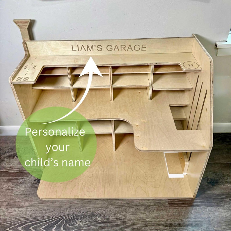 May include: A wooden toy garage with multiple shelves and compartments. The text "LIAM'S GARAGE" is engraved on the top. The image also includes the text "Personalize your child's name".