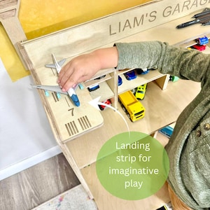 May include: Wooden toy garage with the name "LIAM'S GARAGE" engraved on the top. Features a landing strip with the number 7 and a series of lines. Includes a toy airplane and various toy cars. The text "Landing strip for imaginative play" is also visible.