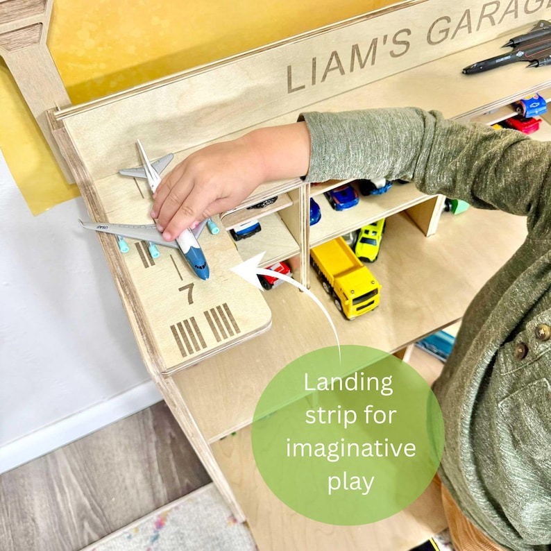 May include: Wooden toy garage with the name "LIAM'S GARAGE" engraved on the top. Features a landing strip with the number 7 and a series of lines. Includes a toy airplane and various toy cars. The text "Landing strip for imaginative play" is also visible.
