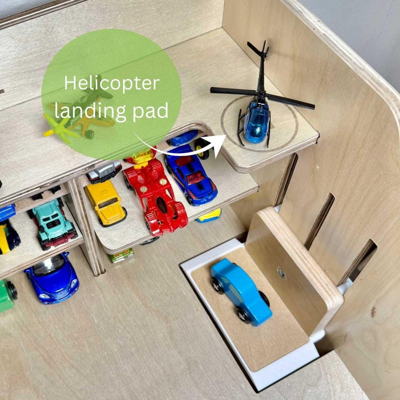 May include: A wooden toy car garage with multiple levels and a helicopter landing pad. The garage is filled with colorful toy cars and a blue helicopter. The text "Helicopter landing pad" is visible.