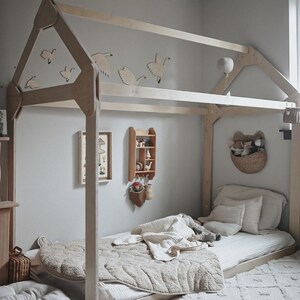 May include: A wooden bed frame shaped like a house with a white duvet and pillows. The bed is decorated with wooden bird cutouts and a small wooden shelf with decorative items.