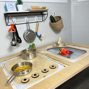 May include: A wooden play kitchen set with a white countertop, two stovetop burners, a sink, and a cabinet. The kitchen set includes a variety of accessories, such as a timer, a wooden rolling pin, a spatula, a strainer, a pot, a pan, and a bowl. The kitchen set is designed for children to play pretend cooking and cleaning.