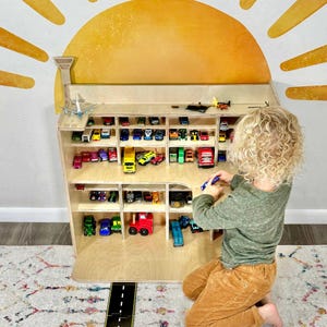 May include: A wooden toy car display shelf filled with various colorful toy cars, trucks, and planes. The shelf has multiple compartments and is designed to resemble a building. A child is playing with a toy car in front of the shelf.