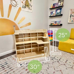 May include: A wooden children's play garage with multiple shelves and a desk area. The garage is 30 inches wide and 25 inches tall. The text "LIAM'S GARAGE" is printed on the top. The garage is made of light-colored wood.