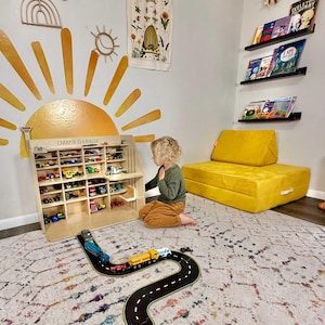 May include: A wooden toy garage labeled "Liam's Garage" filled with toy cars. A child sits nearby, looking at the garage. A black and yellow road mat with toy cars on it is in the foreground. The room has a yellow sofa and wall art.