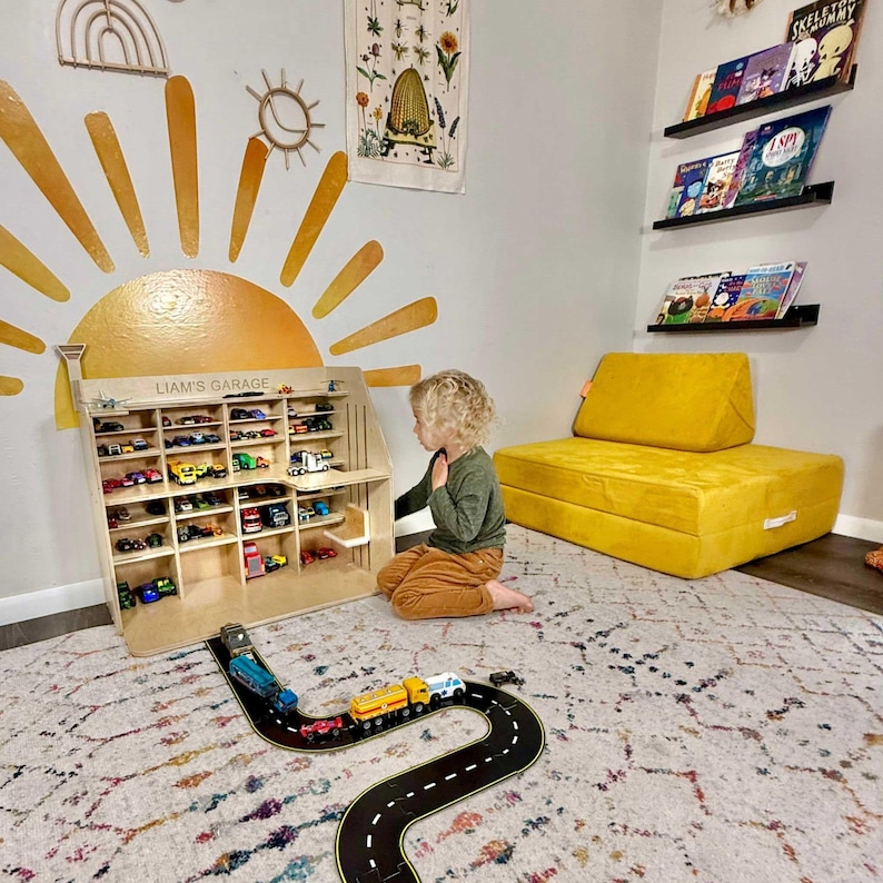 May include: A wooden toy garage labeled "Liam's Garage" filled with toy cars. A child sits nearby, looking at the garage. A black and yellow road mat with toy cars on it is in the foreground. The room has a yellow sofa and wall art.