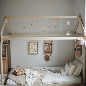 May include: A wooden bed frame with a white canopy and a white and brown blanket on the bed. There are wooden birds and a hot air balloon hanging from the canopy. There is a small wooden shelf with toys on it and a basket with stuffed animals hanging from the bed frame.