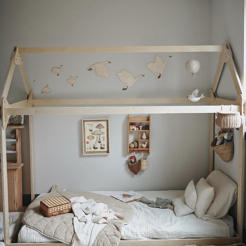 May include: A wooden bed frame with a white canopy and a white and brown blanket on the bed. There are wooden birds and a hot air balloon hanging from the canopy. There is a small wooden shelf with toys on it and a basket with stuffed animals hanging from the bed frame.